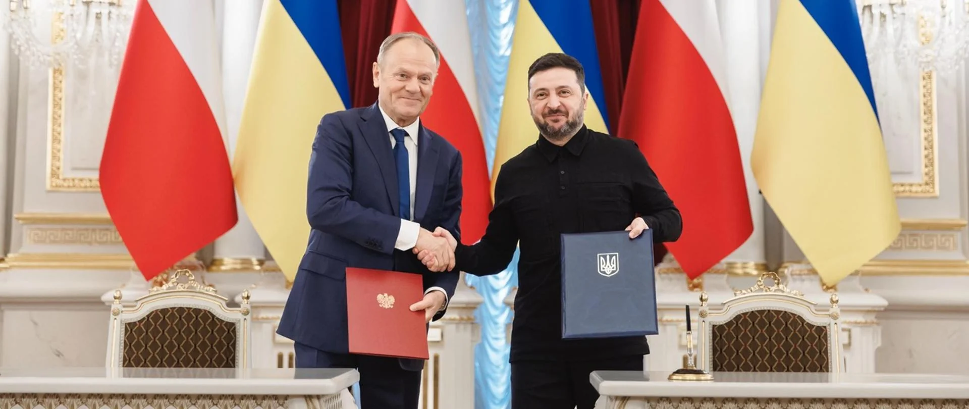 Дональд Туск і Володимир Зеленський тиснуть руки з папками на тлі прапорів